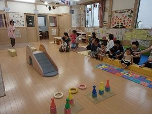 子供の遊びスペースに参加した親子が座り、左奥に立つピンク色のエプロンを着用した女性スタッフがが右手を上げているのを見ている写真