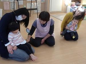 子どもを膝抱っこしている2組の親子が座っている間に、ピンク色のエプロンを着用した女性スタッフが座り、左に座っている子供に中央に12月と書かれた手作りのクリスマスリースの札を見せて顔を覗いている写真