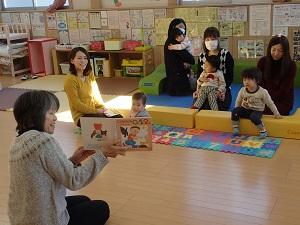 子どもの遊びスペースに4組の親子が座り、左手前に向かいあって座っている女性が絵本を見せながら読み聞かせを行っている写真
