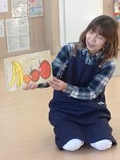 紺色のエプロンを着用した女性スタッフが、バナナやリンゴが描いている果物のイラストを見せながら話しをしている写真