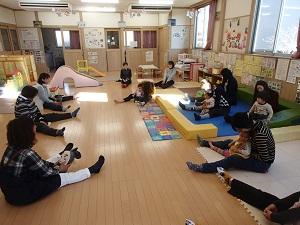 室内で間隔をあけて参加者の母親が座り足を広げた間に子どもを座らせている写真