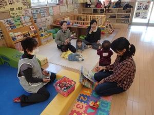 ジョイントマットの上で寝転がっている子供たちやその手前で職員の方が篭におもちゃを戻している写真