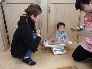 床に置かれた体重計の上に座っている赤ちゃんの横で、女性2人がしゃがんでいる写真