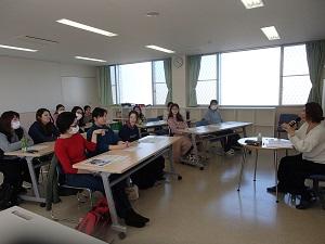 参加者の方々が3人ずつ長机の席に着いて講話に参加しており、赤いセーターを着た参加者と臨床心理士さんが意見交換をしている様子の写真