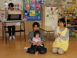 キーボードを弾く先生、黄色いエプロンを付け、手拍子をして歌を歌っている先生、その横でおかあさんの膝に座っている女の子の写真