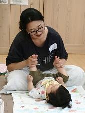 床の上に広げたタオルの上で寝かされた赤ちゃんの両足を持ち、笑顔でベビーマッサージを楽しむ1組の親子の写真