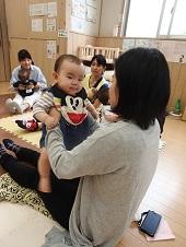 赤ちゃんの両脇を支えたお母さんの膝の上で抱っこされながら、ベビーマッサージを楽しむ1組の親子の写真