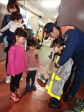 女の子が消防隊の方に消防服を着させてもらっている前で興味深く見る子供の写真