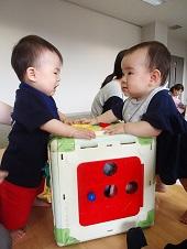 おもちゃが入ったボックスに2名の赤ちゃんが両側に立ち、左側の赤ちゃんが泣きだした写真