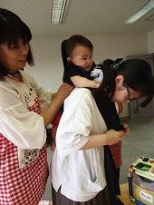 母親が抱っこ紐で赤ちゃんを背負おうとしているのを、後ろのエプロンを着用した女性スタッフが赤ちゃんの脇を持ち手伝っている写真