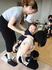 母親が抱っこ紐で赤ちゃんを背負おうとしているのを、後ろの女性が赤ちゃんの脇を持ち手伝っている写真