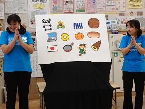 白布にパンダ、黄色の花、パソコン、かぼちゃ、パン、日本の国旗、フライパンなどのイラストが貼られ、両脇に立つ先生が両手を合わせて話をしているパネルシアターの写真
