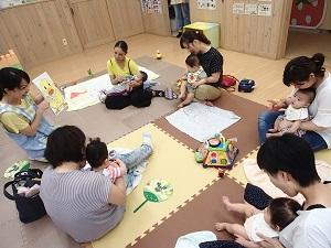 円になって一緒に座っている母親と赤ちゃんに、水色のエプロンを着用した女性スタッフがヒヨコのイラストを見せて話しをしている写真