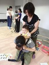 ボーダーシャツを着た母親が、参加者の女性に手伝ってもらいながら抱っこ紐で背負われようとしている赤ちゃんが、母親の髪の毛を触って喜んでいる写真