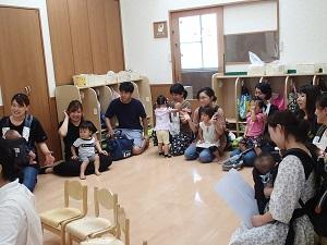 教室の周りに親子連れの参加者の皆さんが座っており、笑顔で2歳児クラスの楽しい英語の時間を見学している写真