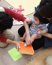 お母さんに抱っこされた赤ちゃんの足型をとるため赤ちゃんの左足を持ち上げ朱肉に押しつけている助産師の写真