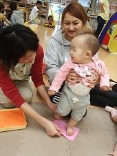 お母さんが赤ちゃんの両脇を支え、立ち上がった状態でピンク色の台紙に左足を乗せ足型をとる助産師さんの写真