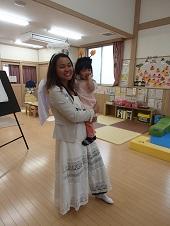 天使のコスチュームを身につけた英語の先生がカチューシャをつけた子供を抱いて立っている写真