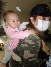 マスクを着け、キャップを被ったお母さんがベージュのおんぶ紐を使って赤ちゃんをおんぶしている写真