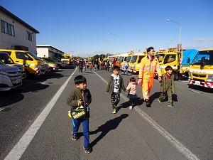 横一列に整列して駐車してあるたくさんの高速道路作業車と手をつないで歩いている子供と作業員の写真