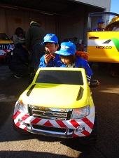 作業車のミニカーに乗っている、青い制服を着た2人の子供の写真