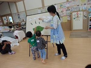ホワイトボードに並べられている青虫とキャベツのシールを幼児2名が保母さんに教えている写真