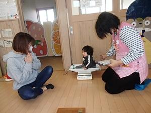 体重計に上手に座る赤ちゃんの横で、お母さんが見守り、保健師さんがメモをしている写真