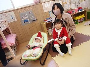 お母さんの膝の上にサンタの格好をした女の子が座り隣にサンタの帽子を被った赤ちゃんがキャリーに寝ころんでいる写真