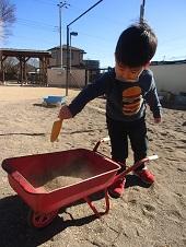 男の子がスコップを使い一輪車に砂を入れている写真