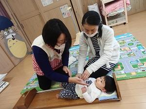 服を着たままで寝転んでいる赤ちゃんの身長を計ろうとしているお母さんと保健師さんの写真