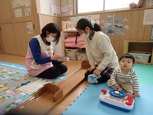 ハンドルのおもちゃで遊んでいる幼児の横で保健師さんと話をしているお母さんの写真