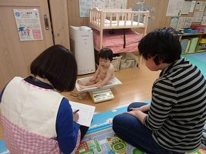 おむつ姿で体重計に座っている赤ちゃんの前でお母さんとメモを取る保健師さんが座っている写真