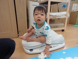 肌着で体重計に乗りながら、左手で床にあるおもちゃを触っている赤ちゃんの写真