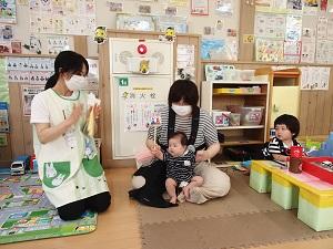 手を叩いている保母さんの隣で赤ちゃんを支えながら立たせているお母さんと、その隣で遊んでいる幼児の写真