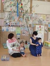 笹飾りの前で小さな男の子が立つのをお母さんが後ろから支え、横に保母さんと男の子が座っている写真