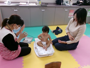 体重を測る乳児と見守る母、助産師