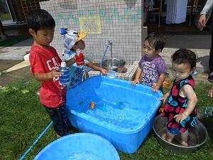 水がためられた水色の四角いたらい、その周りで水遊びを楽しんでいる4人の子供たちの写真