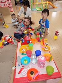 おもちゃのお皿に果物やケーキが入れられ、フォークとセットになって並べられており、その奥に座って遊ぶ男の子と女の子、赤ちゃんとお母さんの写っている写真