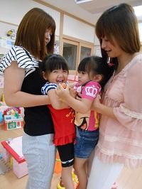 2組のお母さんが背後から女の子がを抱っこして、子ども同士がピッタリくっついており、左側の女の子が笑顔を見せている写真
