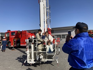 はしご車の箱に乗る家族と、写真を撮ってくれる消防士さん