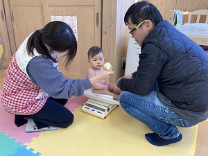 乳児の体重測定をする保健師と見守る父