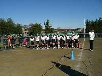 緑色の帽子を被り体操着を着た児童たちが、校庭のスタートラインの内側に立っている写真