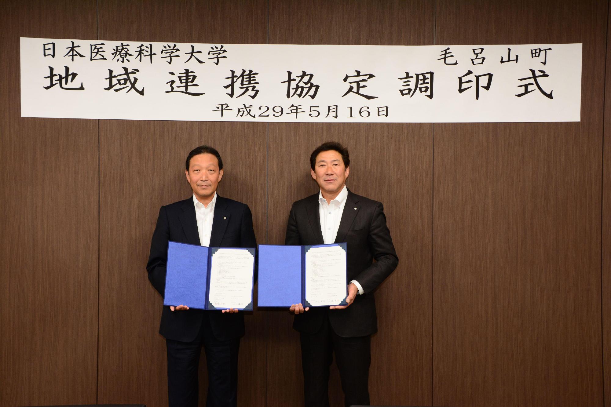「日本医療科学大学地域連携協定調印式」と書かれた幕の前で町長と男性が並んで立ち、協定書を持って写っている写真