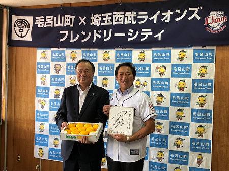 箱に入った柑橘類の果物を持った居郷 肇社長と、サイン色紙を持っている井上 健次町長が並んで写真を撮っている写真