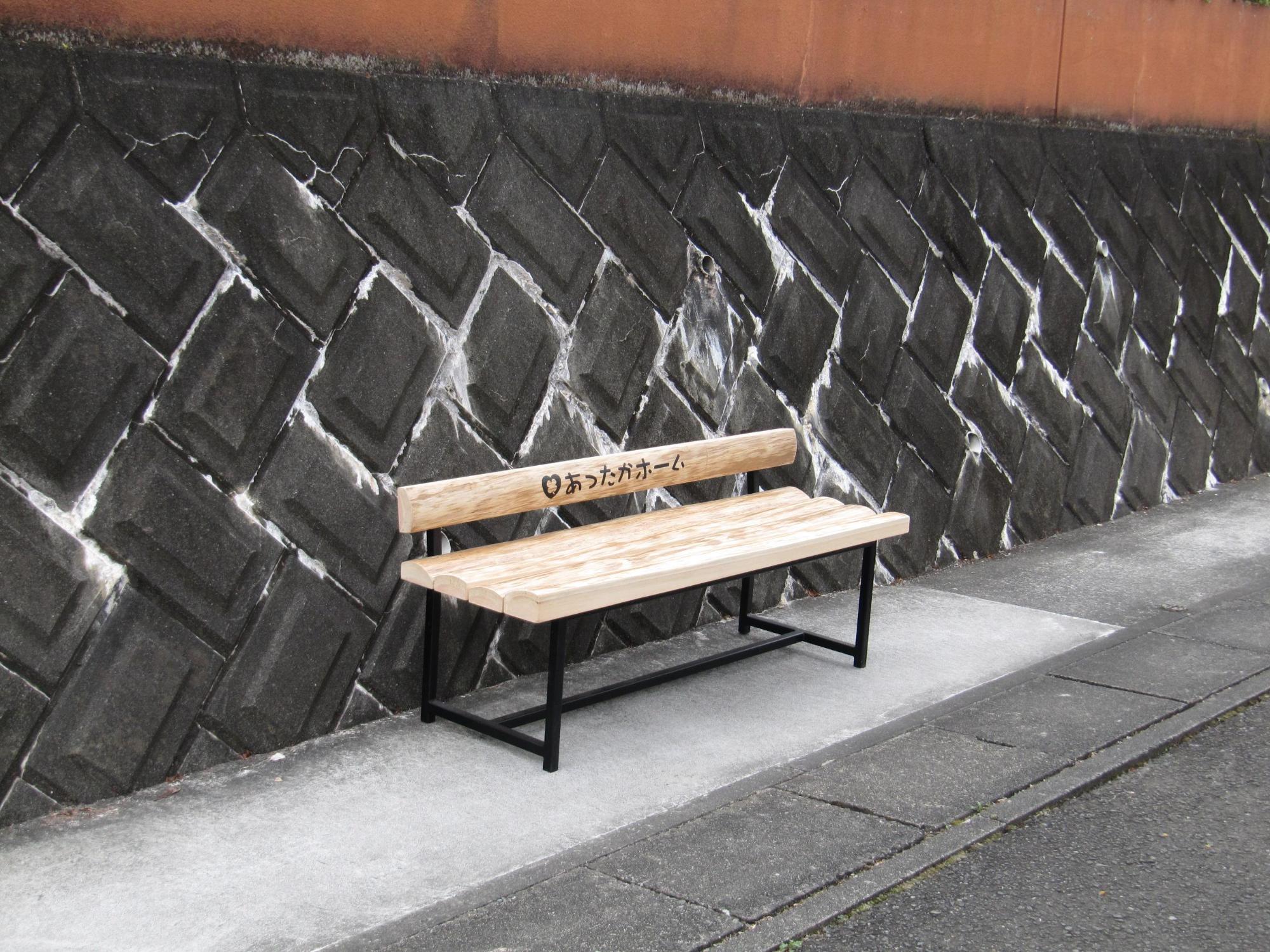 木製のベンチの背もたれに「あったかホーム」と文字の入ったベンチが道路脇に設置されている写真